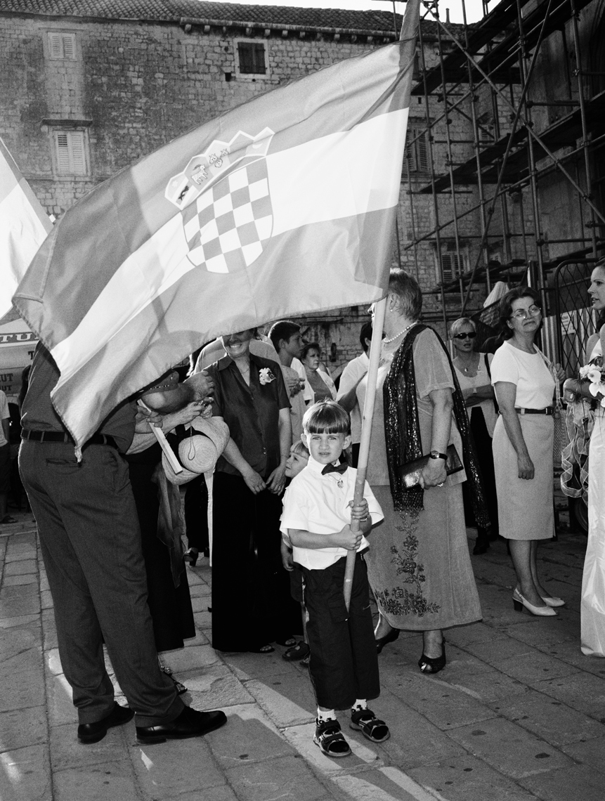 CROATIA, Dalmatian Coast, portrait of a boy holding flag with people in the background B&W